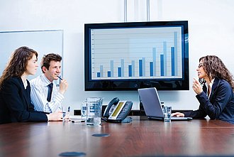 Drei Menschen in Bürokleidung am Konferenztisch vor großem Bildschirm